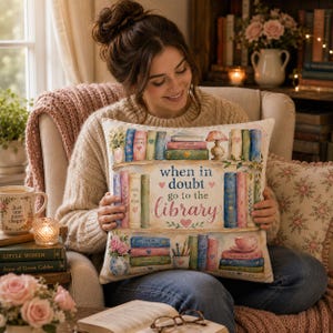 May include: A square throw pillow featuring a watercolor illustration of two wooden bookshelves filled with colorful books, a small lamp, a potted plant, and a teacup. The text in the center reads, when in doubt go to the library.