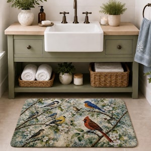 Puede incluir: Una alfombra de baño rectangular con un diseño de pájaros y flores. La alfombra muestra varios pájaros posados en ramas con flores blancas sobre un fondo azul y verde suave. La alfombra está colocada frente a un mueble de baño.