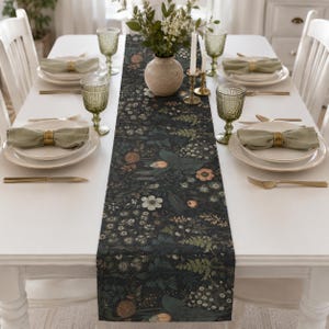 May include: A dark-coloured table runner featuring a botanical print of various flowers, leaves, and ferns in shades of green, white, and muted orange. The runner is centred on a white dining table set with white plates, green glassware, and napkins.