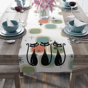 May include: White table runner with a retro design featuring black cats, colorful squares, and stars.