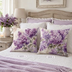 May include: Two square scatter cushions featuring a watercolour-style print of purple lilac clusters on a light background. The cushions are labelled SIDE 1 and SIDE 2, showing different arrangements of the same floral pattern on a white bedspread.