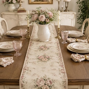 May include: A long rectangular table runner featuring a cream-coloured fabric with a repeating floral pattern of pink roses and green leaves. The runner is centred on a wooden dining table set with white plates, pink textured glasses, and napkins.