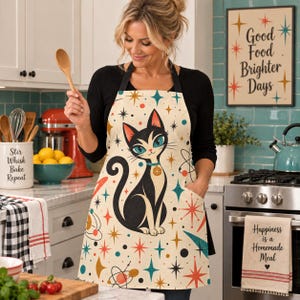May include: A cream-colored apron featuring a black cat illustration with teal eyes and a collar, surrounded by a mid-century modern pattern of colorful stars, planets, and atomic shapes. The apron has black neck and waist ties for securing.