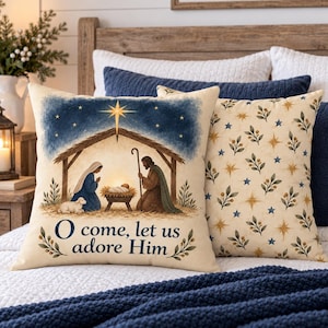 May include: A square throw pillow featuring a nativity scene with a stable, a star, and the text O come, let us adore Him. The scene includes figures in robes, a manger, and a lamb. Beside it is a second pillow with a pattern of stars and leaves.