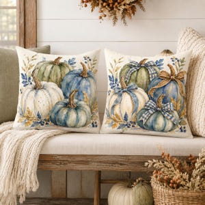 May include: Two square throw pillows featuring a watercolor-style print of pumpkins in shades of blue, green, and cream. The design includes botanical accents, foliage, and decorative bows tied around the stems of the pumpkins on the right pillow.