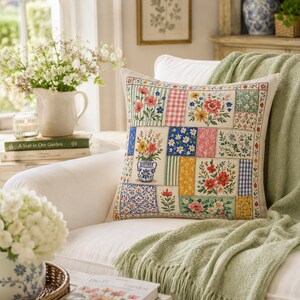 K&ouml;nnte beinhalten: Ein dekoratives quadratisches Kissen mit einem Patchwork-Design mit Blumenmustern in verschiedenen Farben. Das Kissen liegt auf einem wei&szlig;en Sessel, mit einer gr&uuml;nen Decke dar&uuml;ber. Eine Vase mit Blumen und B&uuml;cher befinden sich im Hintergrund.