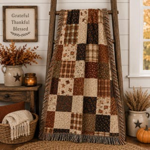 May include: A rectangular throw blanket featuring a patchwork design of various autumn-themed fabrics in shades of brown, cream, orange, and black, including patterns of plaid, small floral prints, and falling leaves, finished with a fringed edge.