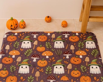 Puede incluir: Una alfombrilla de baño rectangular con un patrón repetitivo de fantasmas blancos con sombreros de bruja verdes, calabazas naranjas y varios hongos marrones y naranjas sobre un fondo morado oscuro con puntos naranjas.
