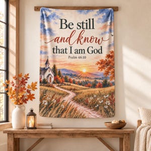 May include: A rectangular wall tapestry featuring a landscape painting of a church, a dirt path, and a sunset. The text reads: Be still and know that I am God Psalm 46:10. The scene includes autumn trees, white flowers, and a blue sky with clouds.