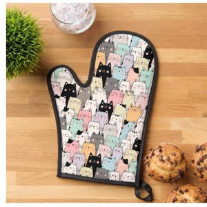 May include: An oven mitt with a black trim and loop, covered in a pattern of cartoon cats in various colors. The cats have different expressions and are set against a light gray background. The mitt is on a wooden surface.