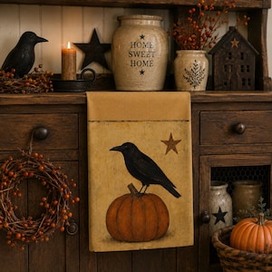 May include: A rectangular fabric wall hanging featuring a mustard yellow top border and a cream-colored lower section. The lower section displays a black crow perched on a round orange pumpkin, accompanied by a brown five-pointed star graphic.