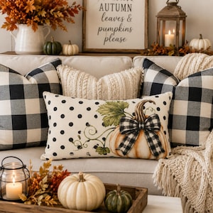 May include: A rectangular throw pillow featuring a cream background with black polka dots and a central illustration of a pumpkin with a green stem and a black and white buffalo check bow, flanked by two square black and white buffalo check pillows.