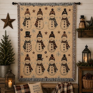 May include: A woven throw blanket featuring a repeating pattern of twelve snowmen on a tan background. Each snowman wears a unique hat and scarf, accented by small stars and snowflakes. The blanket has a fringed edge and is hung on a black rod.