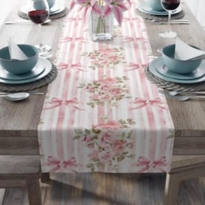May include: A pink and white striped table runner with a floral pattern of pink roses and green leaves. The runner has a watercolour effect and features pink bows.