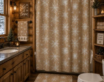 Puede incluir: Una cortina de ducha, una toalla de mano a juego y una alfombrilla de baño rectangular de color tostado, todas con un patrón repetitivo de copos de nieve blancos. La cortina cuelga de una barra negra y la toalla tiene un borde decorativo.