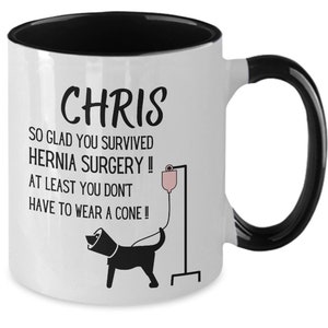 May include: White coffee mug with a black handle and black rim. The mug has a black and white illustration of a dog wearing a cone and a text that reads "CHRIS SO GLAD YOU SURVIVED HERNIA SURGERY!! AT LEAST YOU DON'T HAVE TO WEAR A CONE!!"