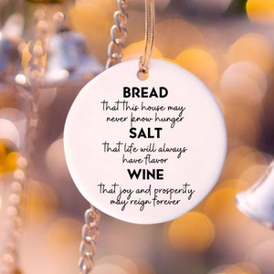 May include: White ceramic ornament with a gold chain. The ornament has black text that reads "BREAD That this house may never know hunger SALT That life will always have flavor WINE That joy and prosperity may reign forever".