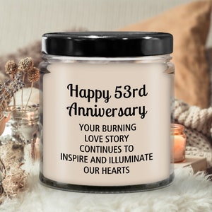 May include: A white candle in a clear glass jar with a black lid. The candle has a label that says "Happy 53rd Anniversary" and "Your burning love story continues to inspire and illuminate our hearts."