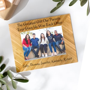 May include: A wooden photo frame with a light brown wood grain pattern. The frame has a photo of five people smiling and sitting on a wall. The text "The Greatest Gift Our Parents Ever Gave Us Was Each Other" is engraved on the top of the frame. The names "Sally, Thomas, Jennifer, Kimberly, Robert" are engraved on the bottom of the frame.