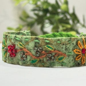 May include: A light green fabric bracelet featuring hand-stitched embroidery of orange flowers, green leaves, and brown stems. The design is accented with small red and metallic beads, with decorative green running stitches along the top and bottom edges.