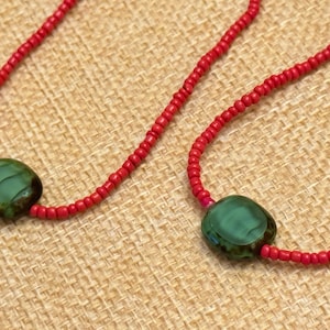 May include: A close-up of a necklace with a red beaded chain. The necklace features two large, oval-shaped, green beads. The beads have a glossy finish and are connected to the red beaded chain. The background is a textured, tan fabric.