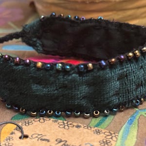 May include: Dark green velvet bracelet with a beaded edge. The bracelet is adorned with small, iridescent beads in shades of gold, blue, and purple. The interior of the bracelet reveals a colourful, patterned fabric. A unique, handcrafted accessory.