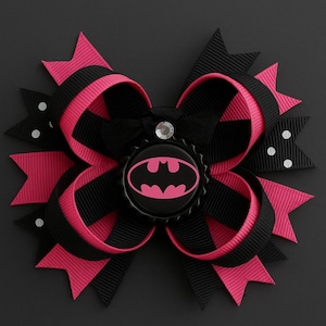Könnte beinhalten: Handgefertigte Haarschleife in Schwarz und Pink mit Batman-Logo in der Mitte. Die Schleife besteht aus Bändern und hat ein mehrschichtiges Design. Sie ist mit einem Strassstein verziert und hat ein auffälliges Aussehen.