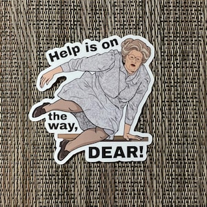 May include: A sticker featuring a cartoon illustration of a woman in a light gray dress, glasses, and a pearl necklace, in a falling pose. The text "Help is on the way, DEAR!" is printed on the sticker.