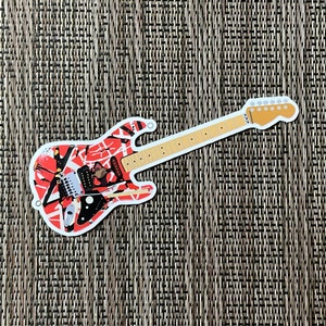 May include: A white electric guitar with a red, black, and white patterned design. The guitar has a black pickguard and a white headstock with black tuning pegs.