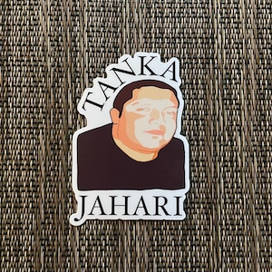 May include: A sticker featuring a portrait of a person with the name "JAHARI" below and the word "TANKA" above. The portrait is in shades of brown and tan, with a black shirt. The background is a textured, woven pattern.