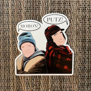 May include: A sticker featuring two cartoon characters, one wearing a blue hat and the other wearing a red and black plaid hat. The character in the blue hat has a speech bubble that says "MORON!" and the character in the red and black plaid hat has a speech bubble that says "PUTZ!"