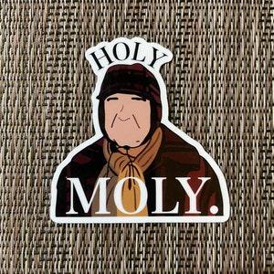 May include: A white sticker with a cartoon illustration of a person wearing a brown hat and a brown scarf. The text "HOLY MOLY." is printed in black letters on the sticker.