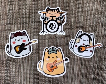 Puede incluir: Cuatro pegatinas de gatos con diferentes instrumentos musicales. Un gato toca la batería, otro la guitarra, otro el bajo y otro el teclado. Las pegatinas son todas blancas con contornos negros y tienen un estilo de dibujos animados.