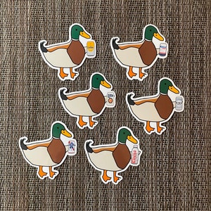 May include: Six colorful stickers of ducks holding different beer cans. The ducks are brown and white with green heads. The beer cans are Miller Lite, Busch Light, Coors Light, Budweiser, and two other brands.