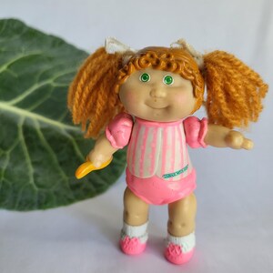 Puede incluir: Una pequeña muñeca de plástico con el pelo rojo en coletas, que lleva un vestido a rayas rosa y blanco y zapatos rosas. La muñeca tiene una cara sonriente con ojos verdes y una tez rosada.