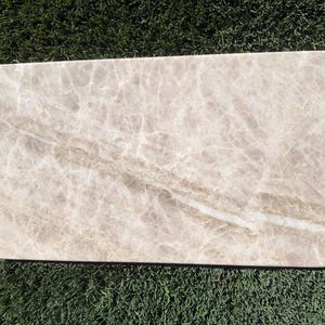 May include: A rectangular slab of polished beige marble featuring a complex network of white veining throughout the surface. The stone displays a smooth, reflective finish with subtle variations in light brown and cream tones across its flat face.