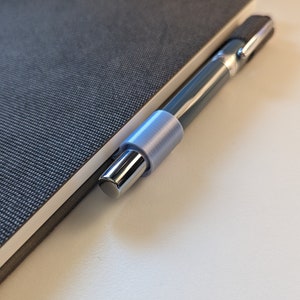 May include: A silver pen with a light blue pen holder is resting on a gray notebook.