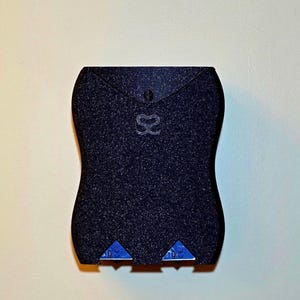 May include: A dark blue, textured wallet with a curved design. The wallet features a small, silver emblem and two blue triangular accents at the bottom. The wallet is closed with a small button.