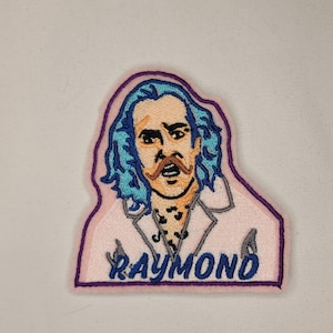 May include: Embroidered patch featuring a cartoon character with blue hair, a moustache, and a goatee. The character is wearing a white shirt with a black and white pattern. The patch has a pink background and a purple border. The text "RAYMOND" is embroidered in blue on the bottom of the patch.