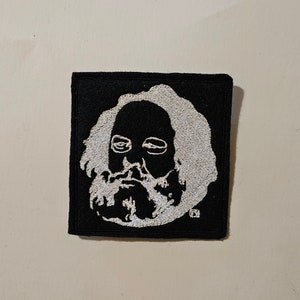 Pode incluir: Patch quadrado preto com um retrato bordado a branco de um homem barbudo. O retrato tem um penteado e barba distintos. O patch é para roupas ou acessórios.