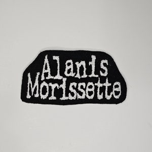 Puede incluir: Parche bordado negro con el nombre "Alanis Morissette" en letras blancas estilizadas. El parche tiene una forma irregular y está diseñado para ser cosido en ropa o accesorios.