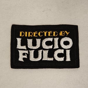 Może przedstawiać: Czarna haftowana łatka z białym tekstem, który brzmi "Directed by Lucio Fulci". Tekst jest w retro stylu czcionki.