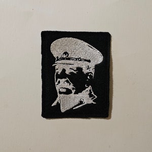 May include: A black and white embroidered patch featuring a portrait of a person wearing a military cap. The person's face is visible, and the patch is on a black background.