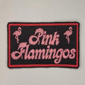 May include: Pink Flamingo embroidered patch with a black background and red border. The text "Pink Flamingos" is in pink.