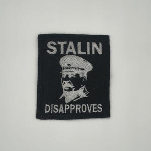 May include: A black square patch with the words "STALIN DISAPPROVES" in white block letters. A white image of a man in a military hat is centered below the word "STALIN". The patch is likely made of fabric.