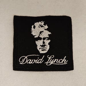 May include: A black embroidered patch featuring a white image of David Lynch and his name in cursive.