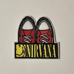 May include: An embroidered patch featuring a pair of red Converse-style trainers with black laces and white soles. The trainers are sitting on top of a black and yellow striped banner with the word "NIRVANA" in yellow letters and a yellow smiley face.