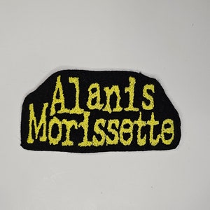Puede incluir: Parche de tela negro con texto bordado en amarillo que dice "Alanis Morissette".