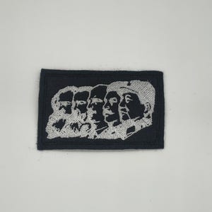 May include: A black rectangular patch featuring a white embroidered design of five profile portraits. The portraits are arranged side by side, with the faces facing right. The patch is likely an emblem or a decorative element.
