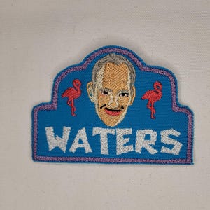 May include: Embroidered patch with a blue background, a portrait, and the word "WATERS" in white. Two red flamingo figures flank the portrait. The patch has a purple border.